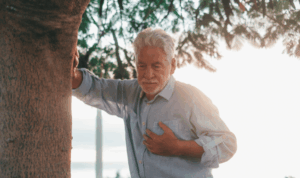 Stariji muškarac se jednom rukom drži za srce, a drugom za drvo u sumrak