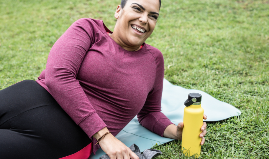 bucmasta žena leži na prostirci na travi nakon treninga, drži bocu u ruci