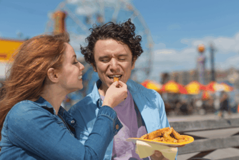 MLadi nasmijani par u zabavnom parku, djevojka mladića hrani prženim krumpirićima