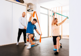 dvje mladih ljudi uz pomoć trenera u dvorani izvodi vježbe s otporom