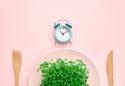 budilica, tanjur pun mikrozelenja i drveni pribor za jelo na ružičastoj podloszi, ilustracija povremenog posta