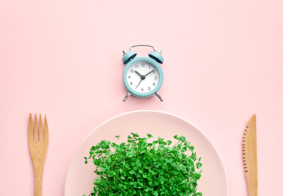 budilica, tanjur pun mikrozelenja i drveni pribor za jelo na ružičastoj podloszi, ilustracija povremenog posta