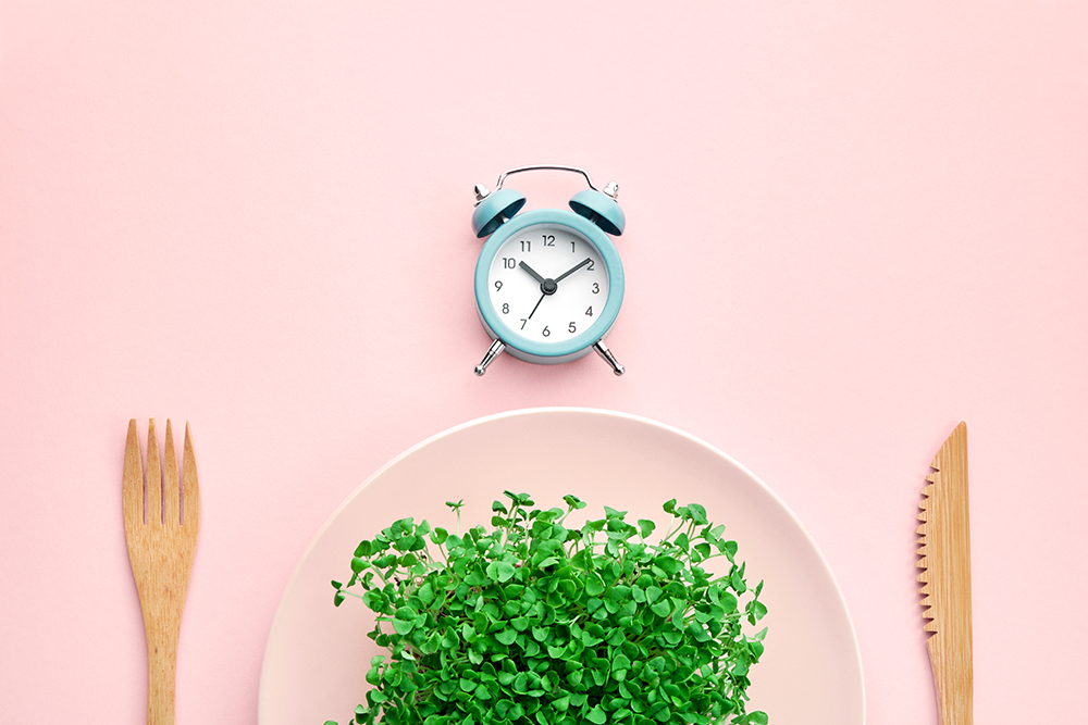 budilica, tanjur pun mikrozelenja i drveni pribor za jelo na ružičastoj podloszi, ilustracija povremenog posta