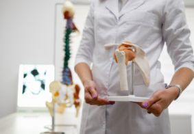 liječnica u ordinaciji pokazje model ljudskog kuka, u pozadini je model kralježnice s kukovima i lubanjom